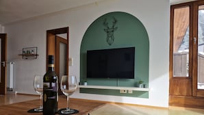 A living room with a wooden table, two wine glasses, a bottle of wine, a mounted television, and a deer head decoration on the wall.