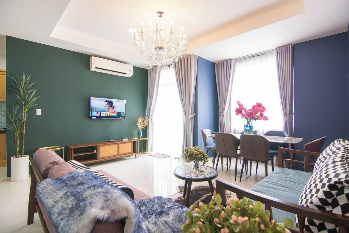 A modern living room with a sofa, a dining area with chairs, a chandelier, and a TV on the wall.