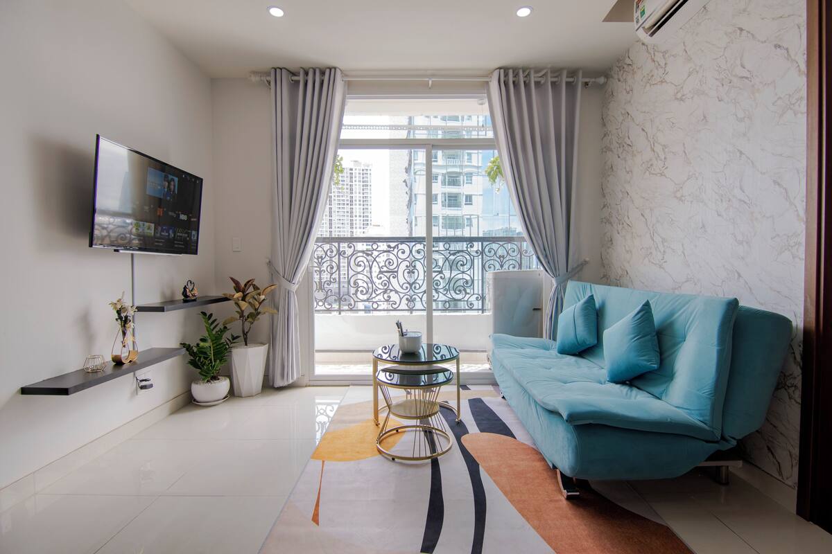 A modern living room with a blue sofa, a flat-screen TV, a small table with plants, and a balcony with a railing.