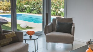 A modern living room with a sofa, armchair, and a small table with a bowl. A large window offers a view of a swimming pool and garden. - Villa 'My Lagozise' with Private Pool, Wi-Fi and Air Conditioning (Pacengo)