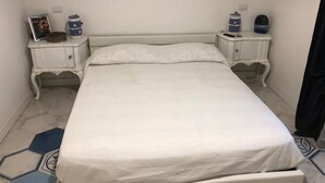 Ein ordentlich bezogenes Bett in einem Zimmer mit weißer Farbgestaltung. Es gibt zwei Nachttische mit dekorativen Accessoires darauf.