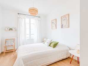 Une chambre à coucher avec un grand lit, une petite table, une chaise, un radiateur et une fenêtre avec des rideaux.