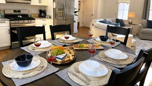 A modern kitchen with white cabinets, a stainless steel refrigerator, and a dining area set with a dark wooden table, woven placemats, and a variety of food and drinks. - National Seashore/Updated/Game Room~Blue Pearl (Eastham)