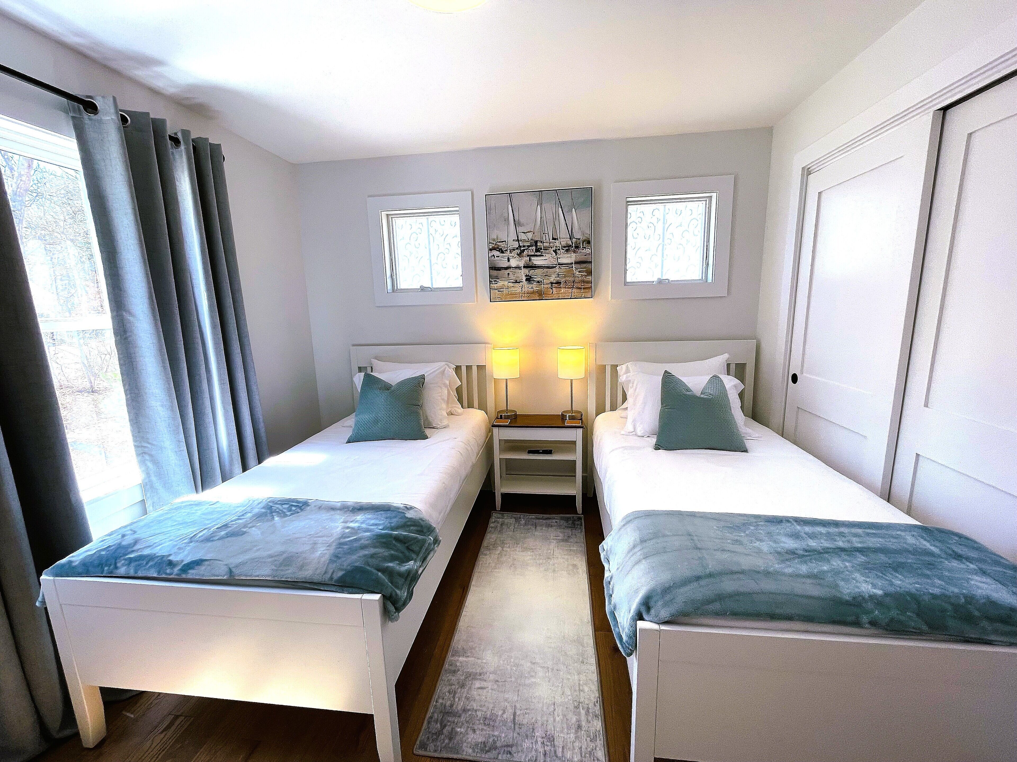 Une chambre double avec deux lits, une table de chevet avec des lampes, une fenêtre avec des rideaux et un tableau au mur.