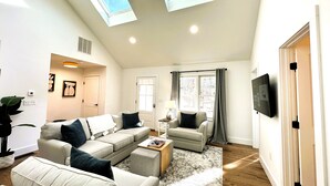 A spacious living room with a high ceiling, skylights, a large sofa set, a coffee table, and a flat-screen TV mounted on the wall. - National Seashore/Updated/Game Room~Blue Pearl (Eastham)