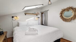 Un dormitorio con una cama grande, dos luces fijadas a la pared, una lámpara en el techo, un espejo con marco de paja y un banco de madera.