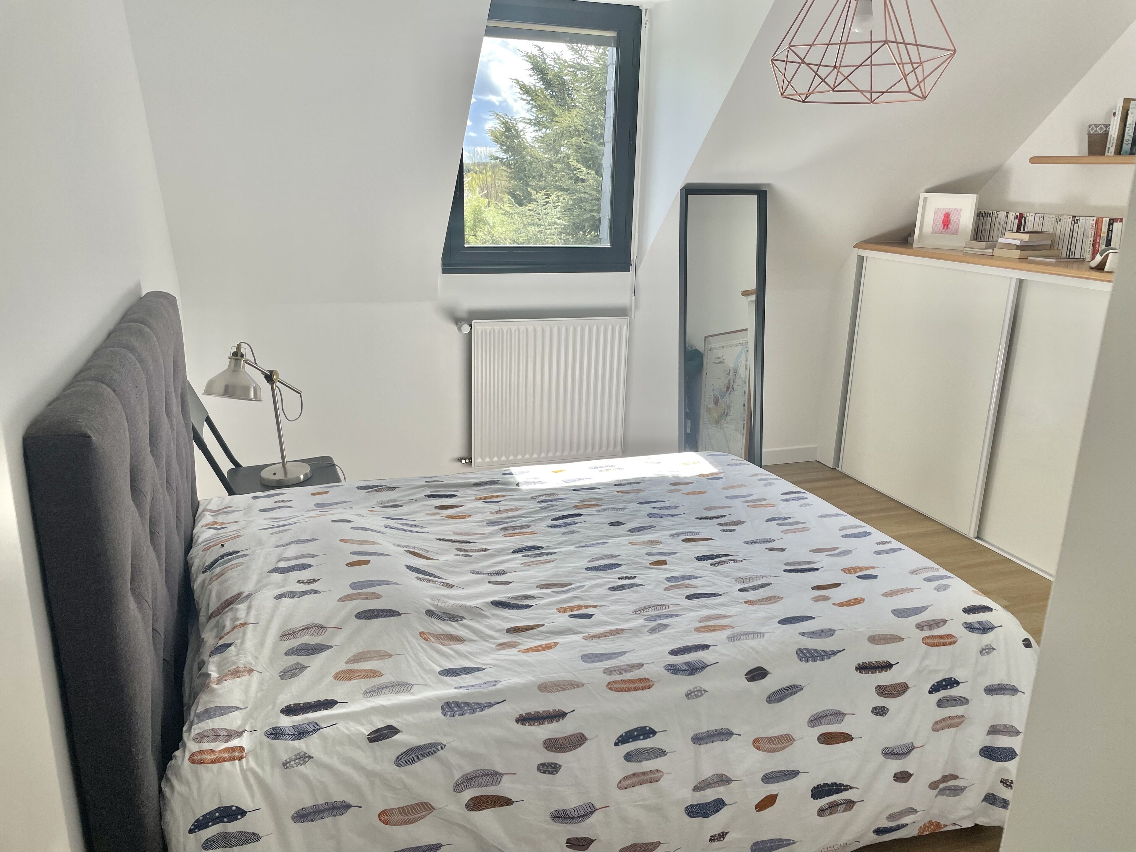 A bedroom with a bed, a window, a radiator, a desk, and a bookshelf.
