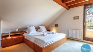 A bedroom with a wooden bed, white bedding, a bedside table with a lamp, and a window with a view. - Le Chapitre - Apartment T4 (Veyrier-du-Lac)