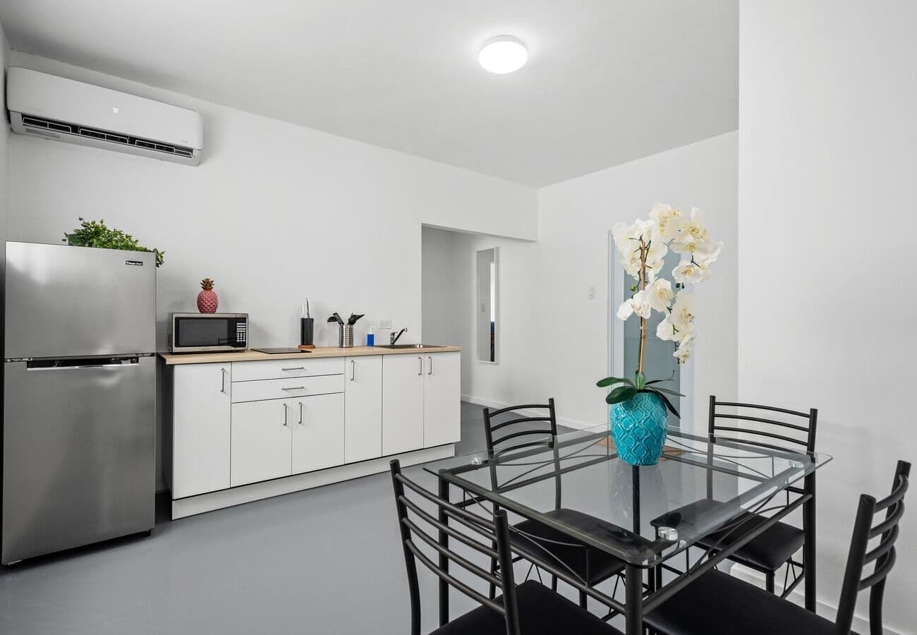 Una cocina moderna con armarios blancos, un refrigerador de acero inoxidable y un microondas. Hay una mesa de comedor con sillas y un florero con flores blancas.