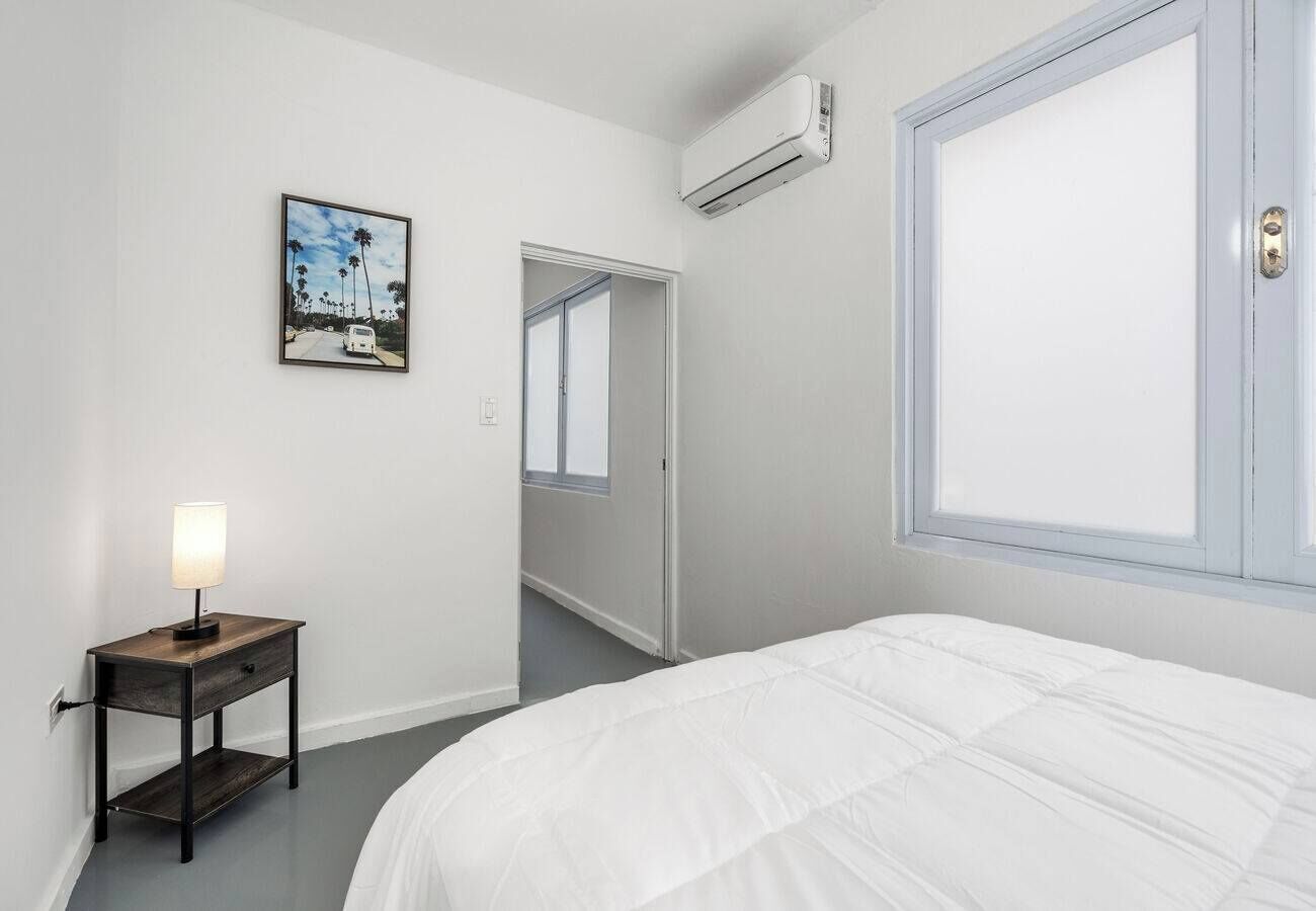 A bedroom with a bed, a nightstand with a lamp, a window, and a door leading to another room.