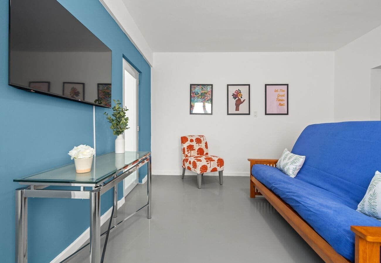 Un salon moderne avec un canapé bleu, une table basse en verre, un fauteuil à motifs rouges et blancs, et un téléviseur fixé au mur.