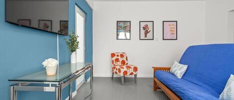Un salon moderne avec un canapé bleu, une table basse en verre, un fauteuil à motifs rouges et blancs, et un téléviseur fixé au mur.
