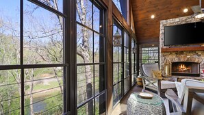 Un rincón exterior acogedor con una chimenea de piedra, ventanas amplias que ofrecen vistas al exterior y un televisor de pantalla plana montado sobre la chimenea.