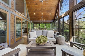 A spacious wooden deck with a sofa and coffee table, large windows offering a view of the outdoors, and a chandelier hanging from the ceiling.