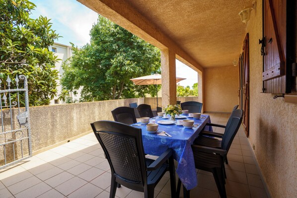 En udendørs terrasse med et spisebord, omgivet af stole, og udsigt til grøn vegetation og bygninger i baggrunden.