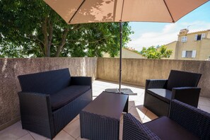 En udendørs patio med fletningsmøbler, herunder en sofa og to stole, under en stor parasol.
