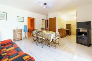 Ein Wohnzimmer mit einer Couch, ein Essbereich mit Tisch und Stühlen, eine Küche inklusive Kühlschrank sowie ein Fernseher.