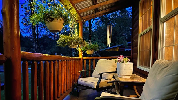 Une véranda en bois avec une balustrade, deux chaises, une table, des jardinières suspendues et une fenêtre.