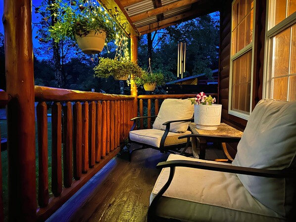 A wooden porch with a railing, two chairs, a table, hanging planters, and a window. - Mossy at Forest Villas: Private Axe Pit, Swing Fire Pit & Pergola Retreat (Marienville)
