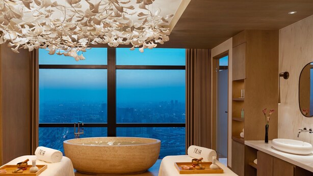 Una moderna spa con una vasca da bagno ampia, due lettini da massaggio, vista sulla città attraverso ampie finestre e un'originale lampada a soffitto.