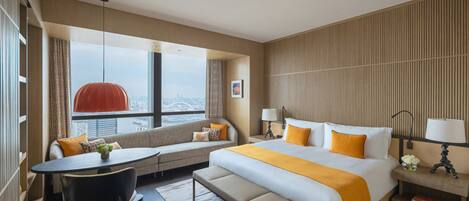 A modern hotel room with a large bed, a sofa, a dining table, and a view of the city.