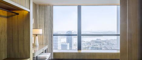A modern hotel room with a large window offering a city view, a desk with a lamp, and a wooden panel on the wall.