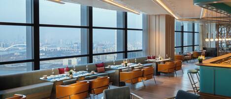 A modern hotel lobby with a panoramic city view, seating areas, and a bar.