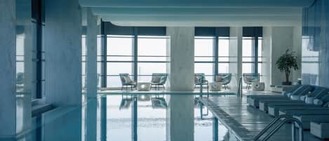 A large indoor swimming pool with clear glass walls, multiple lounge chairs, and a potted plant.