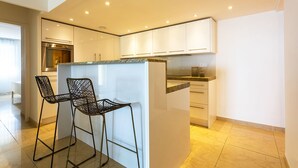 A modern kitchen with white cabinets, a built-in oven, and two black bar stools. - EZE VISTA AP4328 By Riviera Holiday Homes (Eze)