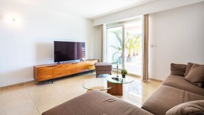 A modern living room with a flat-screen TV, a wooden cabinet, a glass coffee table, a brown sofa, and a sliding glass door leading to a balcony with plants. - EZE VISTA AP4328 By Riviera Holiday Homes (Eze)