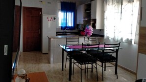 A compact kitchen with a dining area, a refrigerator with magnets, a window with blue curtains, and a vase with flowers on the table. - Cozy villa with large private pool and complete barbecue (El Mirador del Penedès)