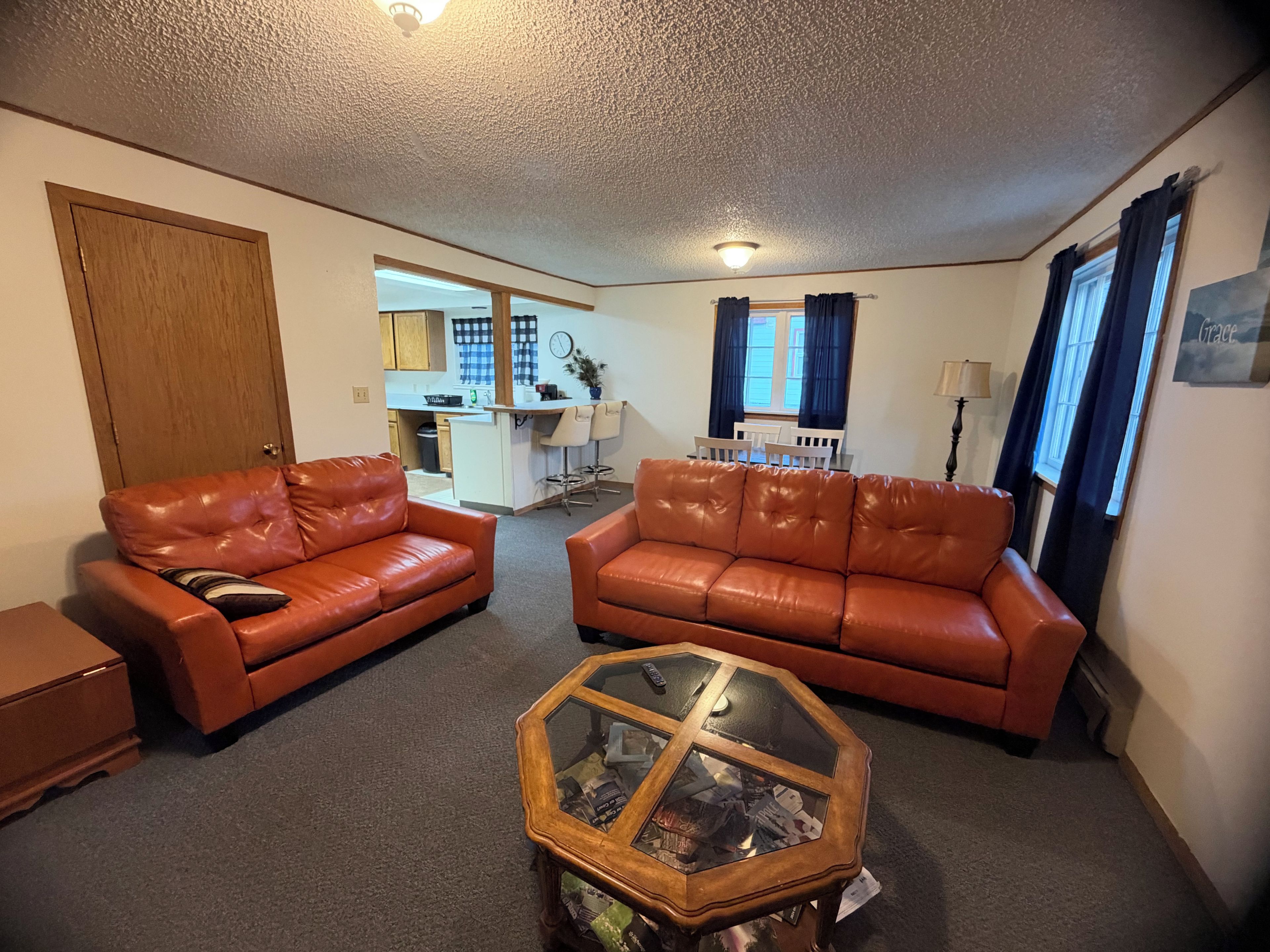 A living room with two leather sofas, a glass coffee table, and a kitchen in the background.