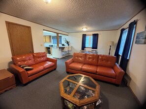 A living room with two leather sofas, a glass coffee table, and a kitchen in the background.