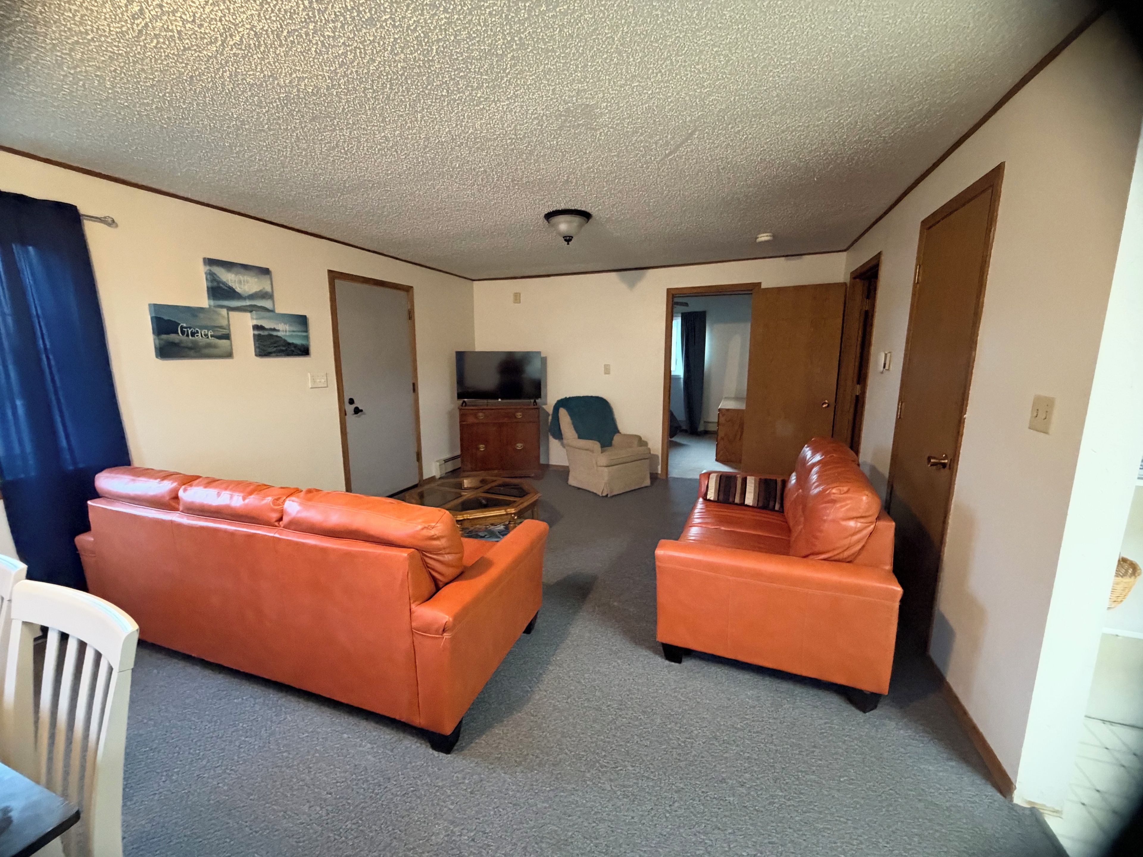 Una sala de estar con dos sofás naranja, una mesa de centro de vidrio, un televisor sobre un mueble de madera y una puerta que da a otra habitación.