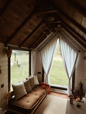 Ein gemütliches Zimmer mit Holzbalkendecke, einer Couch mit Kissen, einem Fenster mit Vorhängen und Blick ins Freie.