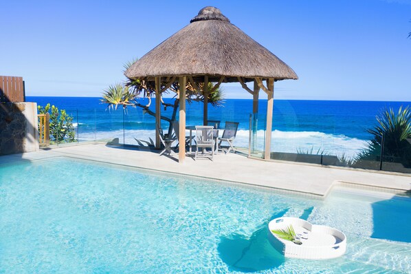 A clear blue pool with a thatched-roof gazebo and lounge chairs by the ocean.