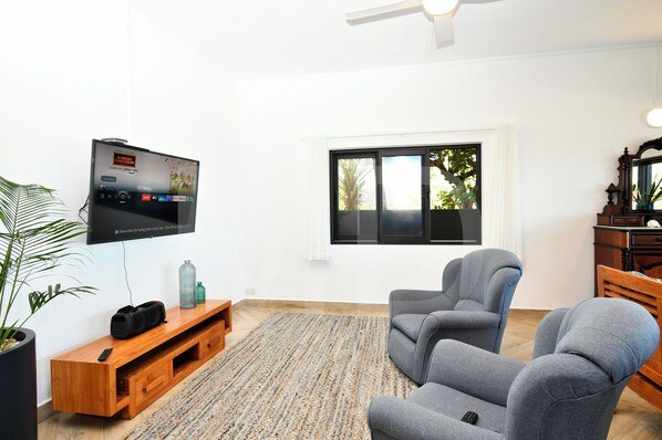 A modern living room with a flat-screen TV, grey armchairs, a wooden entertainment unit, and a large window with a view of trees.