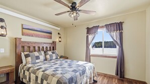 A bedroom with a wooden bed, a ceiling fan, a window with curtains, and a painting on the wall. - Family-Friendly Old West Cabin Near YNP (Island Park)