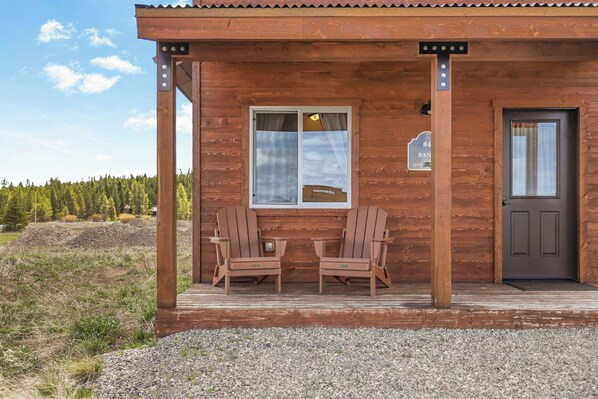 Une cabane en bois avec deux fauteuils Adirondack à l’extérieur, une porte et une fenêtre sur laquelle est accroché un panneau.