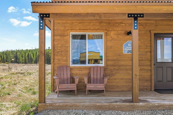 A wooden cabin with a porch, two Adirondack chairs, a door, and a window.