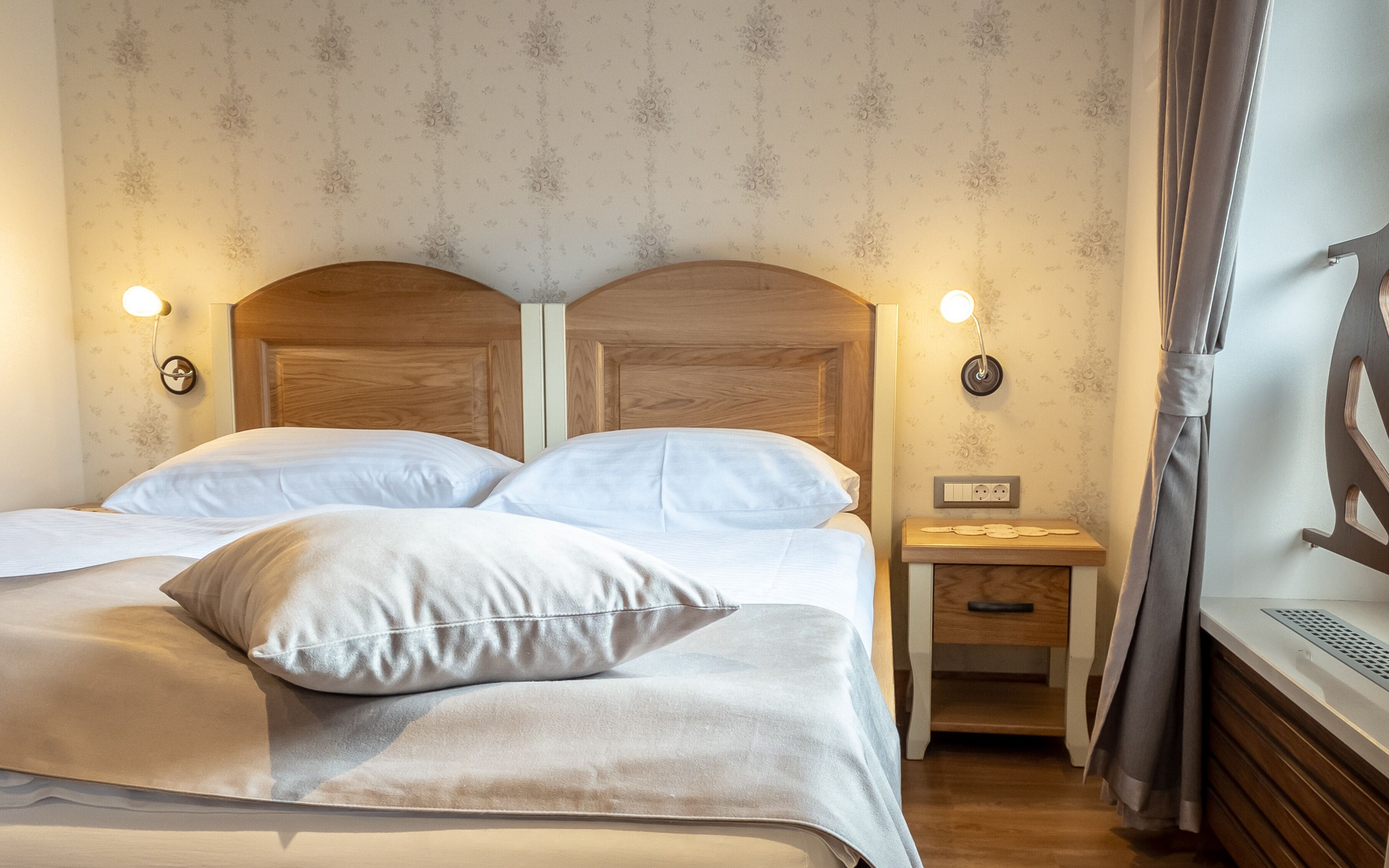 A double bed with white linens, two bedside tables with lamps, a nightstand with a lamp, and a wooden headboard.