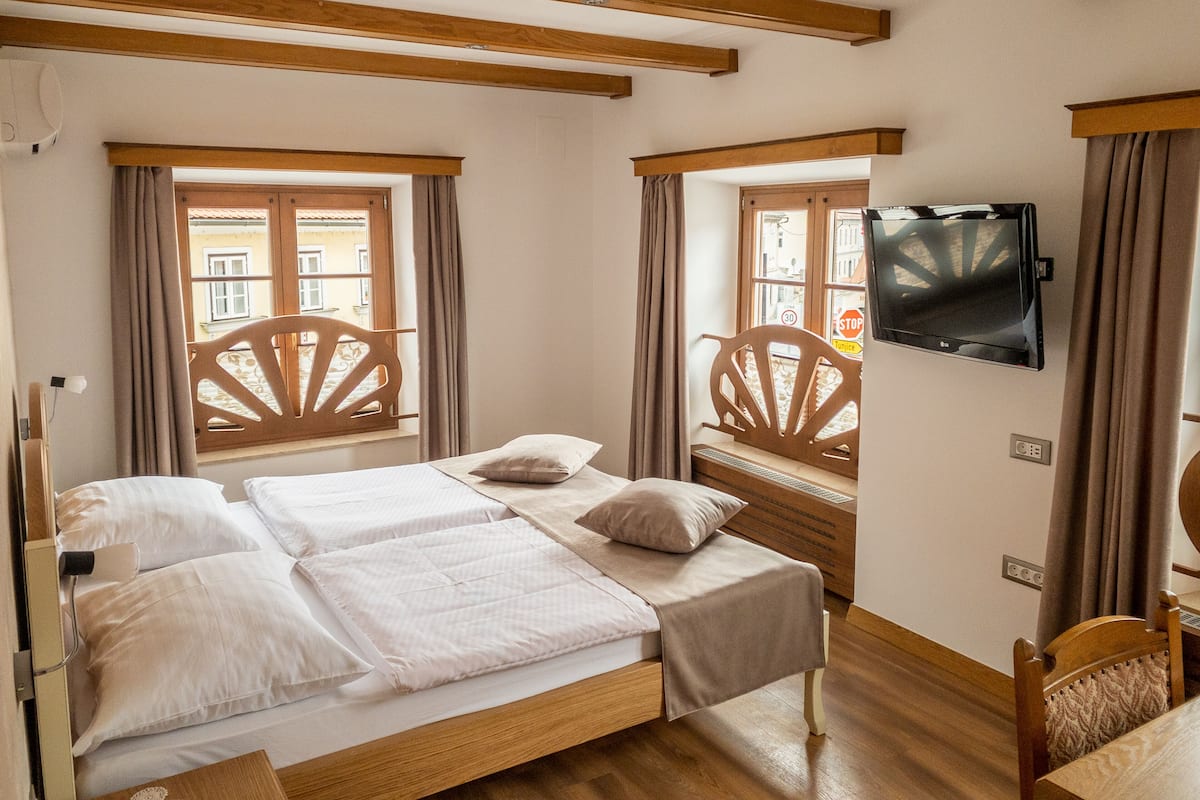 A room with a wooden bed, a television on the wall, and a window with a view of buildings.