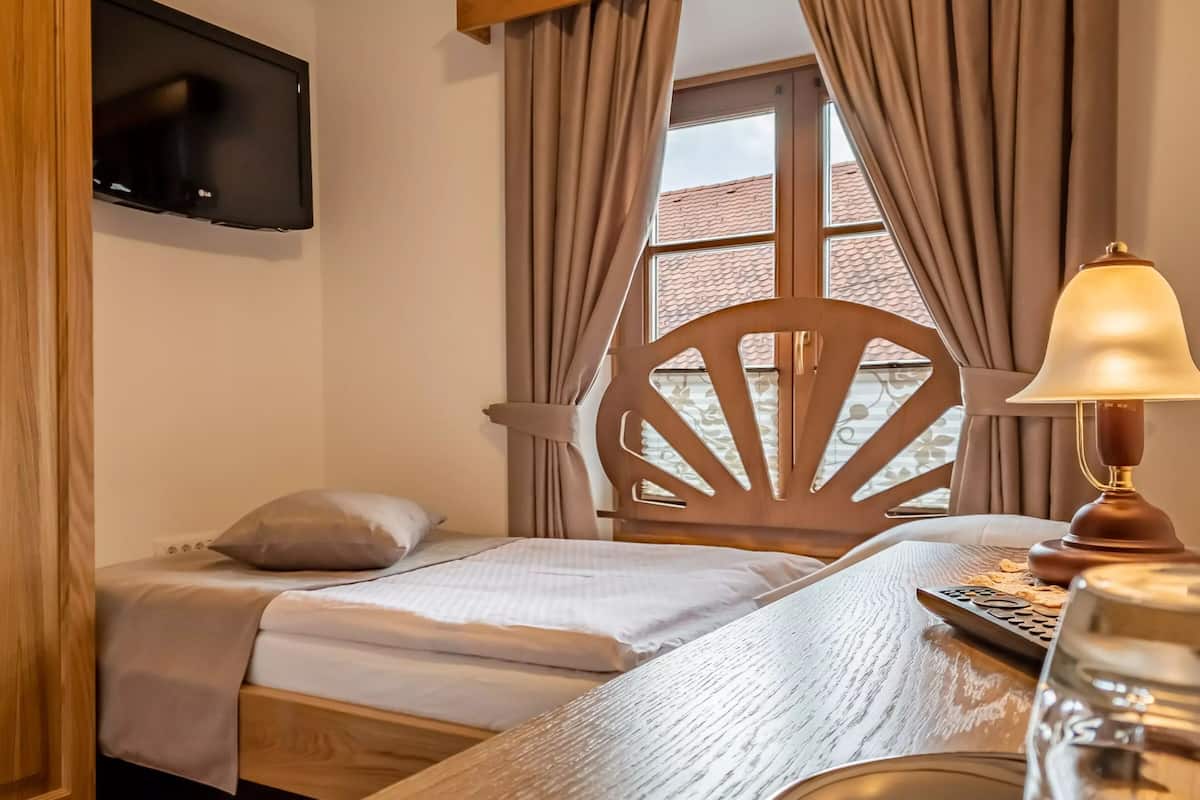 A hotel room with a bed, a wooden chair, a TV mounted on the wall, and a lamp on the nightstand.