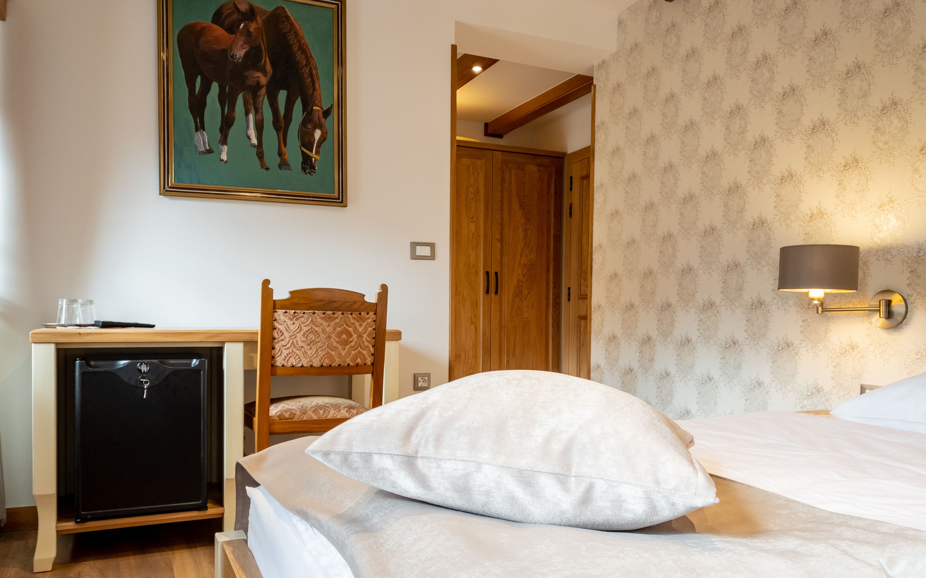 Habitación de hotel con una cama, un escritorio con refrigerador, una silla, un armario de madera y una lámpara de pared.