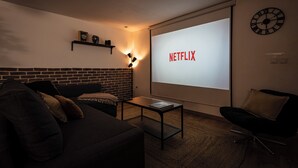 Tunnelmallinen olohuone, jossa on tiiliseinä, musta sohva, lehtien päällä oleva sohvapöytä ja suuri näyttö, jossa näkyy teksti ”NETFLIX”.