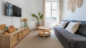 Moderni olohuone, jossa on litteänäyttöinen televisio, harmaa sohva, puinen sivupöytä, pyöreä puinen sohvapöytä ja ruukussa oleva kasvi.