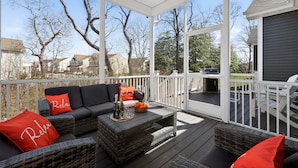 A covered outdoor deck with a sofa, cushions, and a table set with bottles and oranges. - Arbor-Lyn * 4BR Luxury House, Sleeps 10 (Rehoboth Beach)