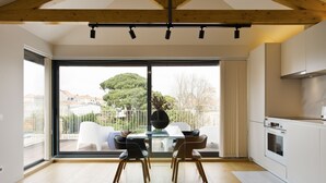 A modern kitchen with a large sliding glass door leading to a balcony, a dining area with a glass table and chairs, and a view of a residential area with trees and houses.