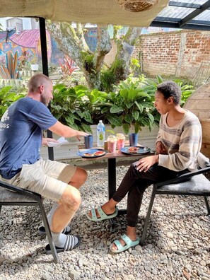 Two people seated at an outdoor table with drinks and food, surrounded by greenery and a brick wall.