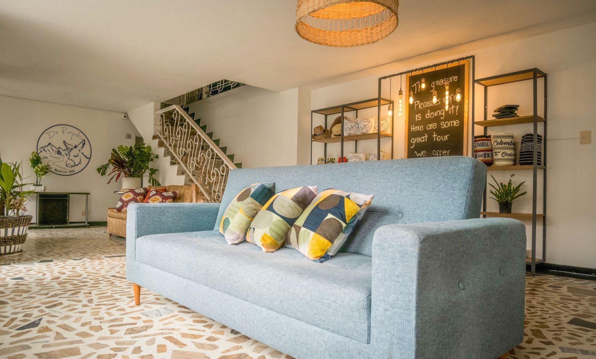 A living room with a blue sofa, patterned cushions, a staircase, a bookshelf with hats, and a wall with a sign.